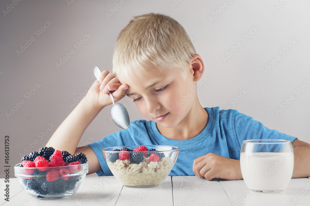 Sad child does not want to eat porridge with berries. Little boy eats without appetite. The concept of a healthy breakfast for children, porridge with raspberries and blackberries, a glass of yogurt. Sad child does not want to eat porridge with berries. Little boy eats without appetite. The concept of a healthy breakfast for children, porridge with raspberries and blackberries, a glass of yogurt.