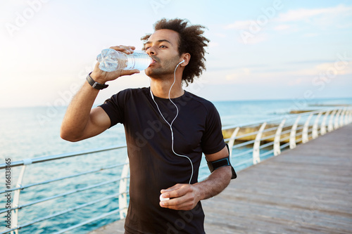 Stylish Afro-American male runner drinking water out of plastic bottle after cardio workout, wearing white earphones Fototapet