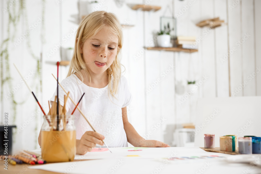 Shot of adorable blonde girl with freckles biting her tongue because of inspiration while painting. Girl with blond hair sitting at the room filled with morning light and wearing white clothes. Shot of adorable blonde girl with freckles biting her tongue because of inspiration while painting. Girl with blond hair sitting at the room filled with morning light and wearing white clothes.