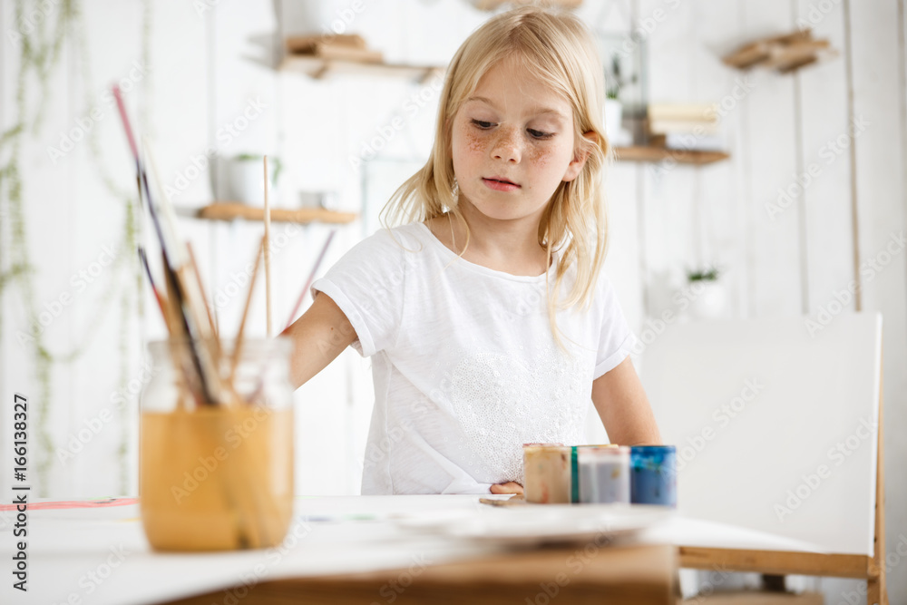 Happy, playful and cute blonde girl with freckles and beautiful blue eyes dressed in white t-shirt biting brush, thinking about watercolour picture she is drawing. Children, art and happiness concept Happy, playful and cute blonde girl with freckles and beautiful blue eyes dressed in white t-shirt biting brush, thinking about watercolour picture she is drawing. Children, art and happiness concept
