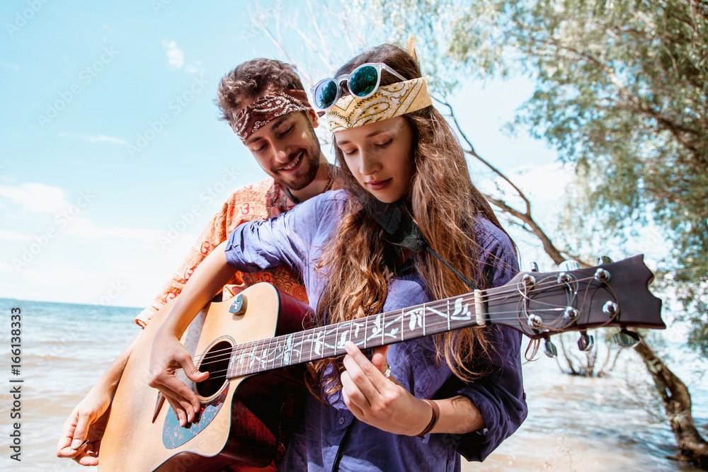 Beautiful young couple in love in hippie style with the guitar resting on the beach in summer. A Girl holding a guitar, man teaches how to play his girlfriend on the seafront
