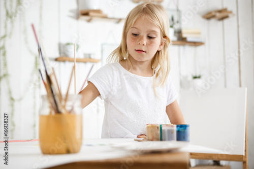 Happy, playful and cute blonde girl with freckles and beautiful blue eyes dressed in white t-shirt biting brush, thinking about watercolour picture she is drawing. Children, art and happiness concept
