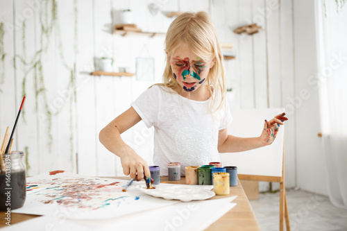 Little blonde girl busy and concentrated on mixing paint on palette. Cute and charming female kid with paint on face captured by inspiration.