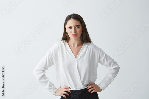 Portrait of displeased young businesswoman with arms akimbo looking at camera over white background.
