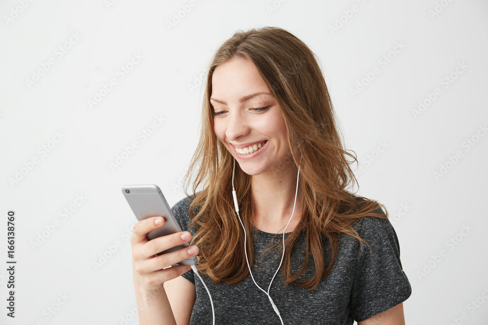 Happy cheerful girl smiling looking at phone screen listening to streaming music over white background. Happy cheerful girl smiling looking at phone screen listening to streaming music over white background.