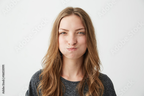 Portrait of young pretty offended girl with funny face looking at camera frowning over white background. © Cookie Studio