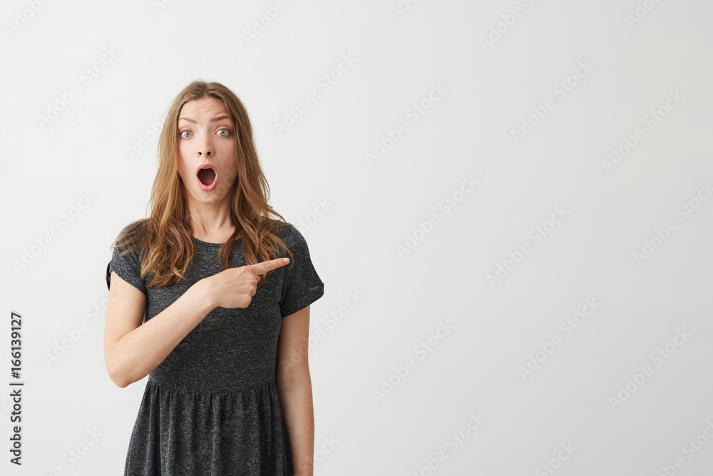 Surprised young positive pretty girl smiling looking at camera pointing finger in side over white background. Surprised young positive pretty girl smiling looking at camera pointing finger in side over white background.