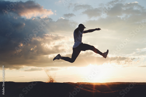 Silhouette of a strong athlete jumping over the hills. Man at sunset in nature is engaged in fitness. Intentional dark colors