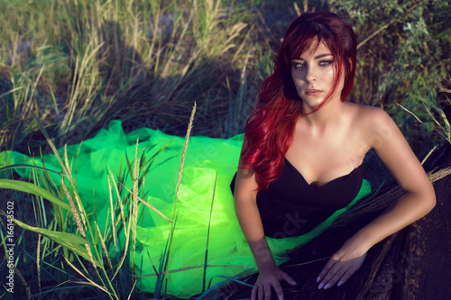 Portrait of young beautiful red haired woman in black corset and long tail green veiling skirt leaning on the shabby upside down wooden boat in the middle of the field with dry grass. Copy-space