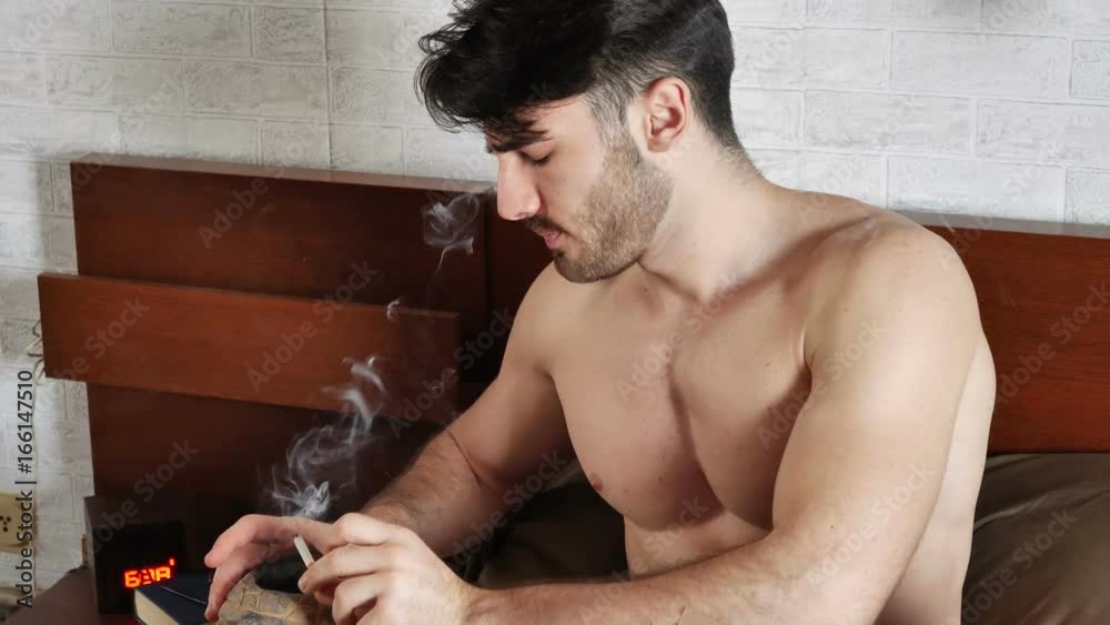 Topless good-looking fit young man smoking cigarette in bed at home before going to sleep. Dangerous situation. Slow motion.