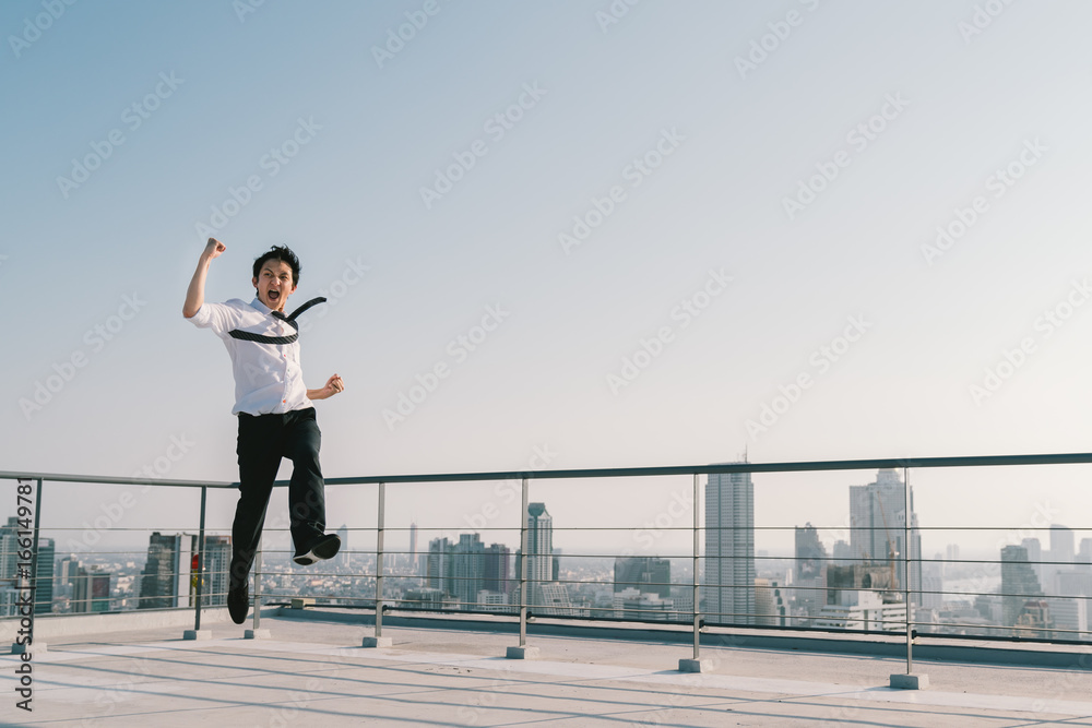Young handsome Asian businessman jumping high, celebrate success winning pose on building rooftop. Work, job, or successful business concept. Cityscape background with copy space on sunny blue sky