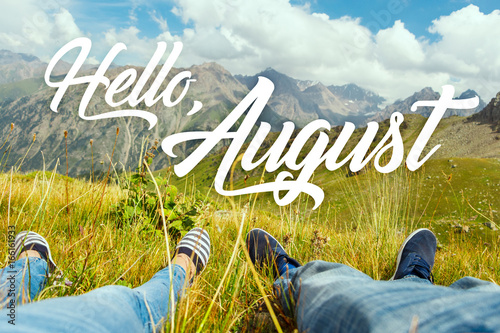 Hello august text with couple's legs together on view of the green valley and high mountains. Nature calendar background. Travel, vacation, holiday, unity with nature, adventure concept.