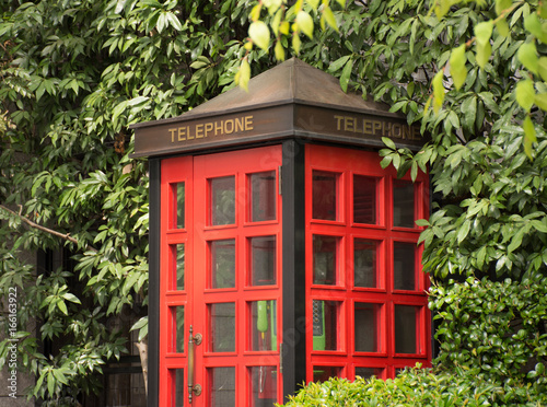 Red Telephone Booth with multiple small window panes and a green telephone. Lush green trees and shrubs surround the telephone booth. 