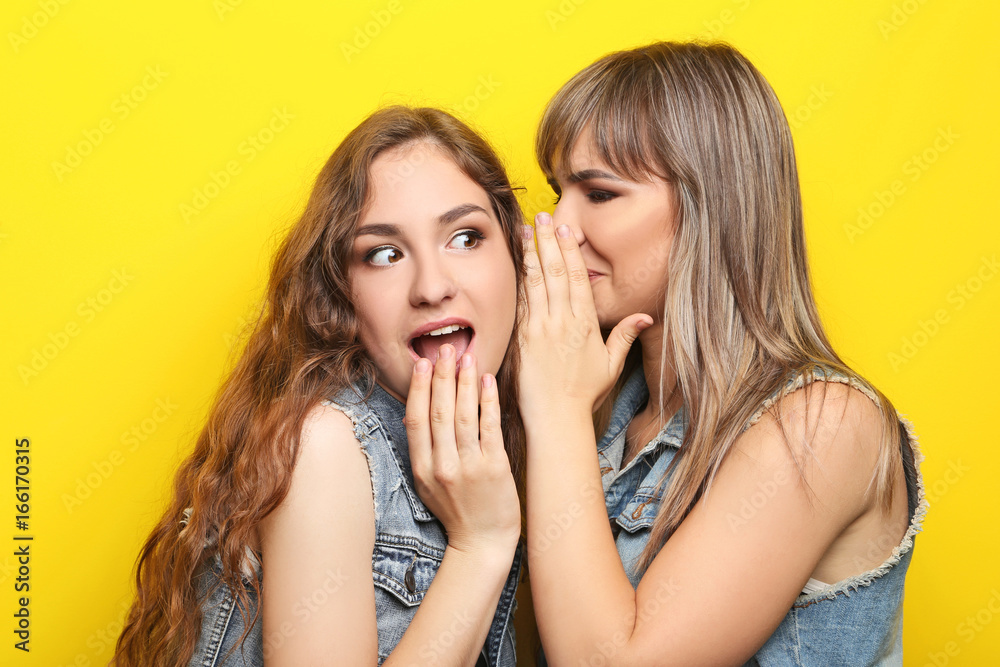 Two young woman whispering a secret on yellow background