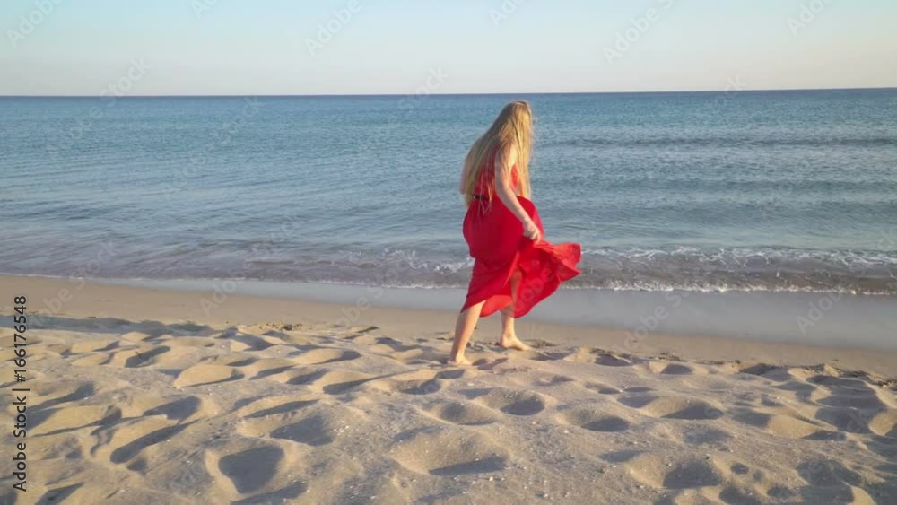 Young barefoot girl in long red dress walking along surf line on sandy beach. Teen girl with long blonde hair whirling and smiling on evening sea shore. Gimbal shot. Young barefoot girl in long red dress walking along surf line on sandy beach. Teen girl with long blonde hair whirling and smiling on evening sea shore. Gimbal shot.