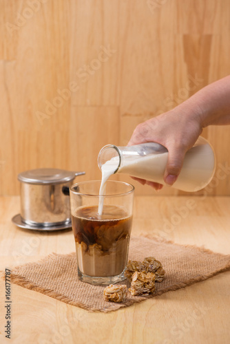 Pouring milk in to glass of coffee on a wooden table.