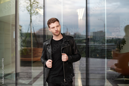 Cool young European male with bristle and stylish haircut standing outside office building with glass door, having confident look, adjusting his trendy black leather jacket. People and style concept