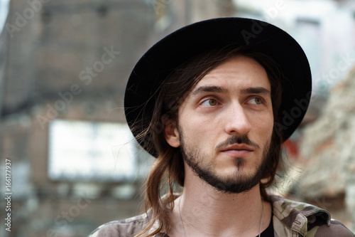 Headshot of fashionable young bearded European traveler in stylish hat having walk in old town during summer holidays. Close up outdoor shot of handsome unshaven hipster standing in urban setting