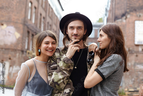 Attractive young European man wearing trendy hat and camouflage jacket relaxing in urban setting with his two girlfriends, enjoying good company. Three cheerful teenage friends having fun outdoor