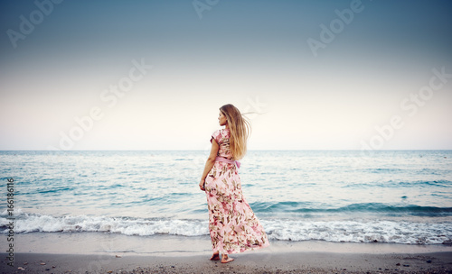 Young female in long dress standing on tropical coast enjoying ocean wind and sunset.