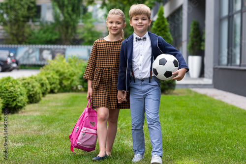 beautiful happy schoolboy and schoolgirl with backpack and soccer ball smiling at camera near school