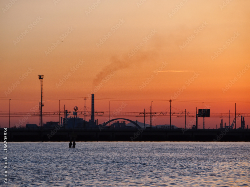 orange sunset sky on the venice lagoon showing railway bridge to mestre and industrial chimneys with smoke and dockside cranes