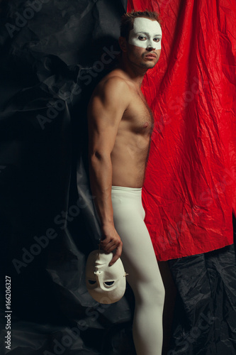 Arty portrait of circus performer in tights holding venetian mask (volto bianco), posing over black and red cloths. Muscular body and perfect tan. Studio shot
