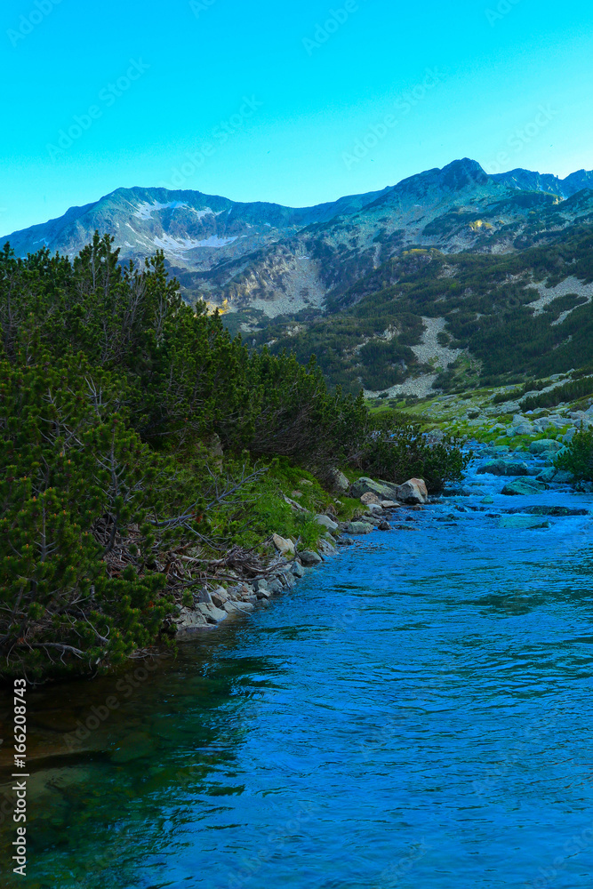 Beautiful view on the high green mountains peaks and a mountain river, blue sky background. Mountain hiking paradise landscape, A stream flows down the rocks, no people.
