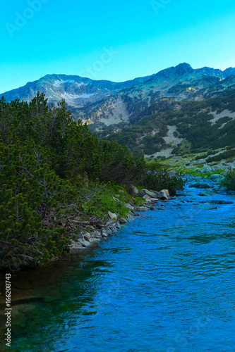 Beautiful view on the high green mountains peaks and a mountain river, blue sky background. Mountain hiking paradise landscape, A stream flows down the rocks, no people.