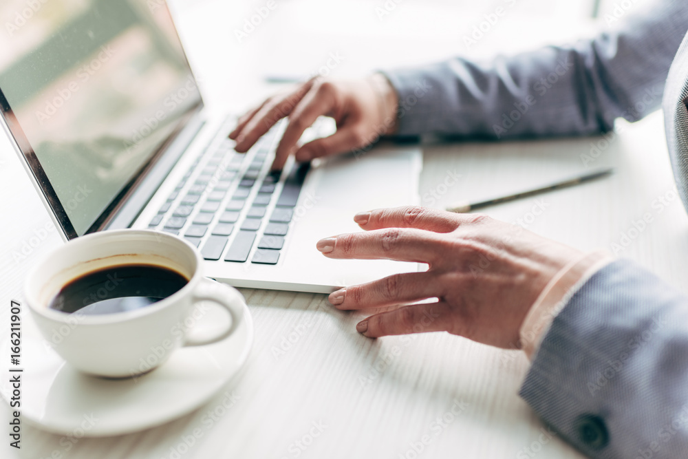 cropped view of businesswoman typing on laptop with cup of coffee on foreground cropped view of businesswoman typing on laptop with cup of coffee on foreground