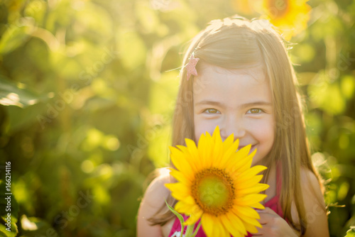 Beauty little girl with sunflower enjoying nature and laughing on summer sunflower field. Sunflare, sunbeams, glow sun
