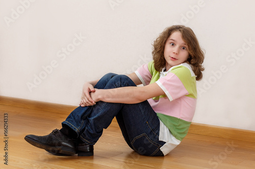 The eleven-year-old boy with long hair sits on a floor and astonishment looks in a lens