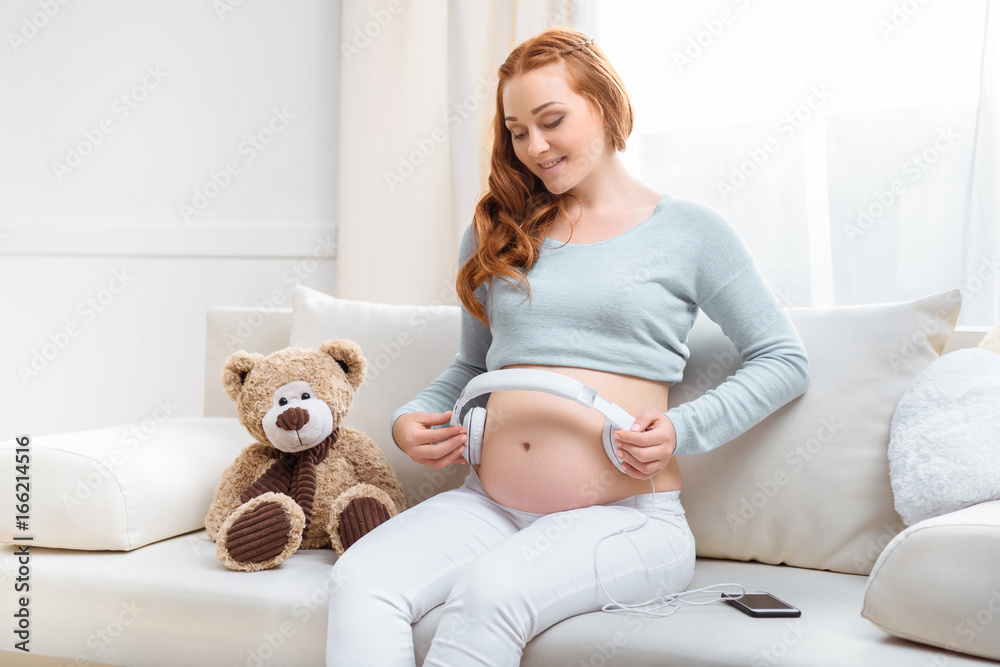 charming redhead pregnant woman with headphones on her belly sitting on  sofa with teddy bear, Stock image