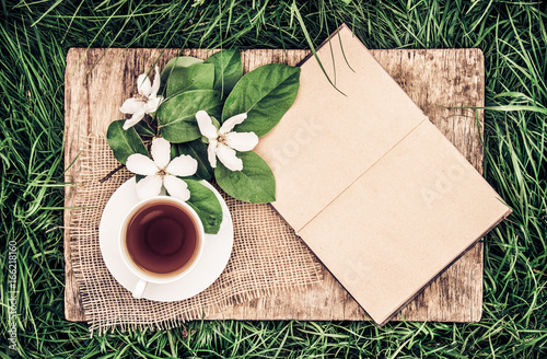 An open book with empty pages, a cup of hot tea and a quince flower. Vintage tinting. Breakfast in the garden. Tea on the green grass.