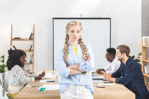 portrait of upset businesswoman with arms crossed looking at camera and colleagues working behind in office © LIGHTFIELD STUDIOS