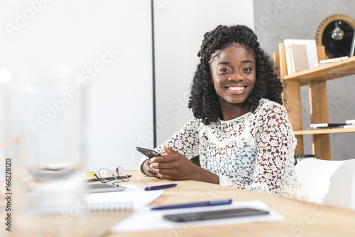 portrait of smiling african american businesswoman with smartphone in hands looking at camera while sitting at workplace