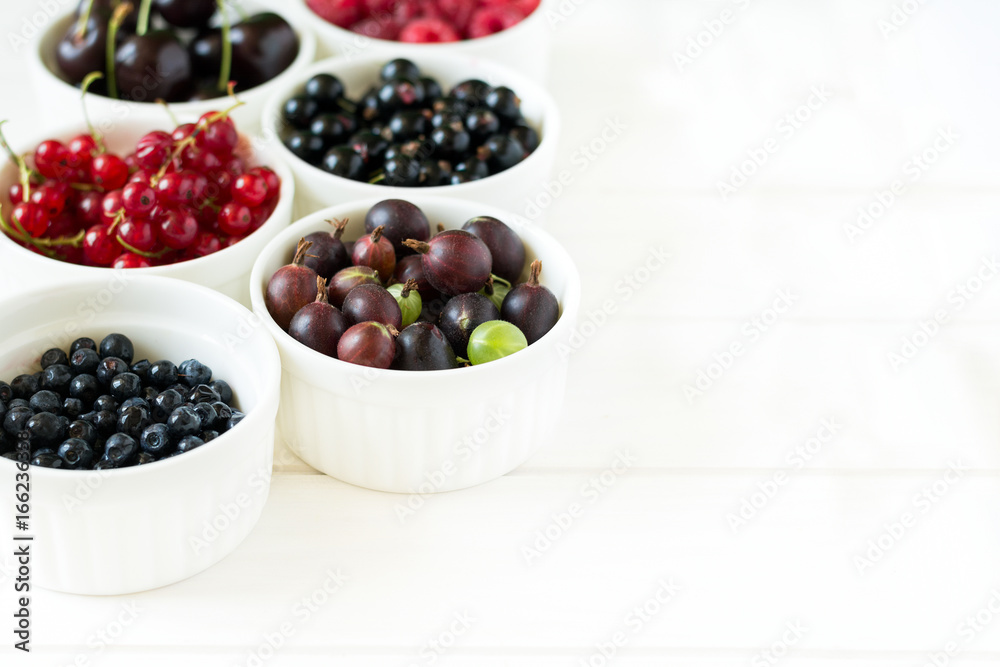 Bowls with fresh ripe gooseberry, red currant, black currant, raspberry, blueberry and cherry on white wooden table Bowls with fresh ripe gooseberry, red currant, black currant, raspberry, blueberry and cherry on white wooden table