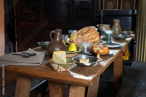 Rustic old wooden dining table in a medieval cottage, laid with bread, meat, cheese and fruit, and antique pottery and pewter