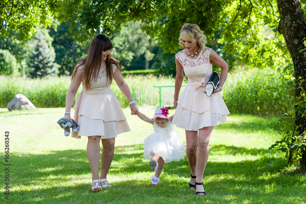 Obraz premium Funny baby girl in white dress is playing with mother and godmother in the garden at christening