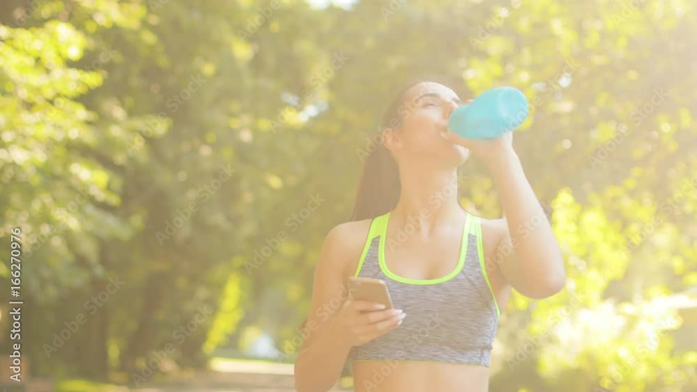 Young woman drinking water after running and using smart phone on the green park background. Young woman drinking water after running and using smart phone on the green park background.