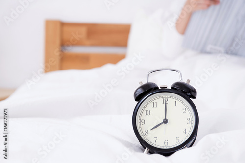 Beautiful asian woman laying and reading book in bed sleep behind alarm clock, time concept.