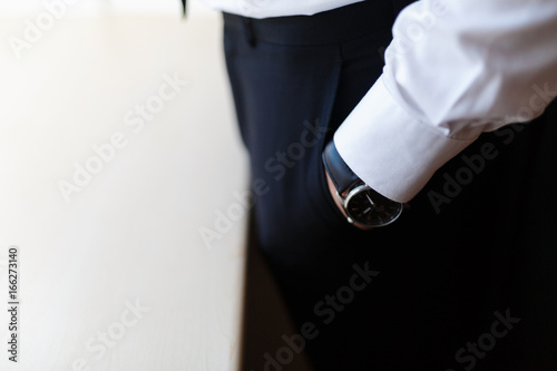Businessman in white shirt holding hands with watches in the pockets of black trousers in an hotel room before an important meeting. Serious handsome man in casual white shirt, black tie, black pants.