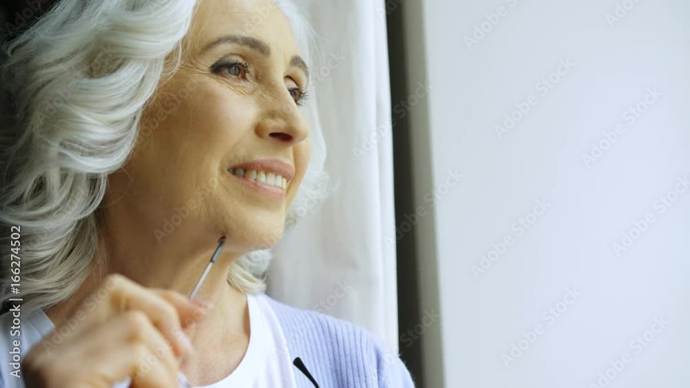 Close up portrait of old woman with eyeglasses and grey hair smiling and looking throught the window outside. Indoor shot.