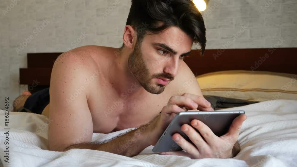 Handsome young man at home in his bedroom using tablet computer while lying on his bed