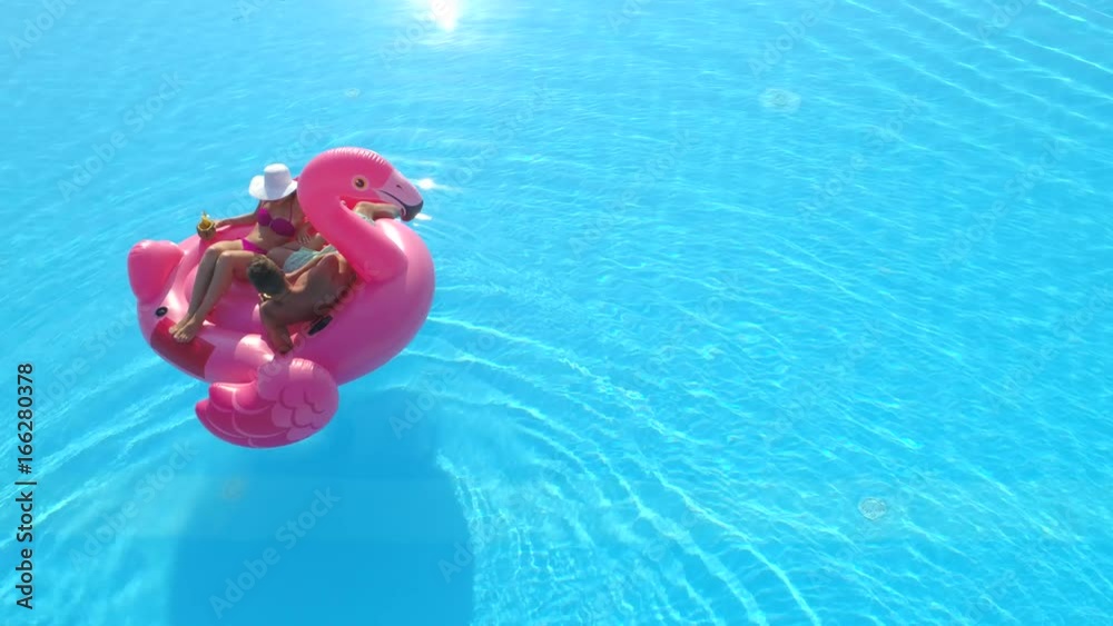 AERIAL: Young couple drinking cocktails and relaxing while lying on fun inflatable flamingo floatie. Sexy couple in swimsuit sipping drinks and enjoying on pink flamingo floating on sunny pool water
