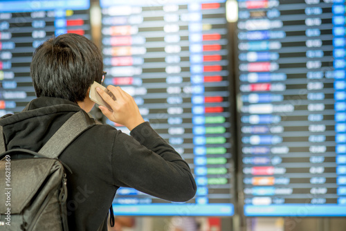 Young asian man with backpack bag checking his flight from the flight information board and using smartphone at airport terminal