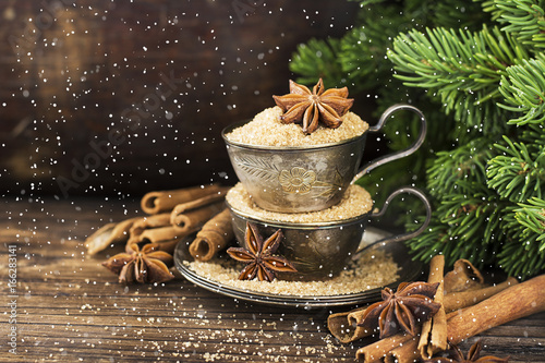 Melchior vintage cups with cane sugar, anise stars, Indian Indian cinnamon sticks on an aged wooden background surrounded by spruce spruce branches. with imitation of falling snow