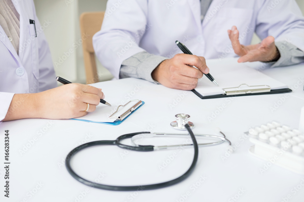 Two doctors discussing patient notes in an office pointing to a clipboard with paperwork as they make a diagnosis or decide on treatment Two doctors discussing patient notes in an office pointing to a clipboard with paperwork as they make a diagnosis or decide on treatment