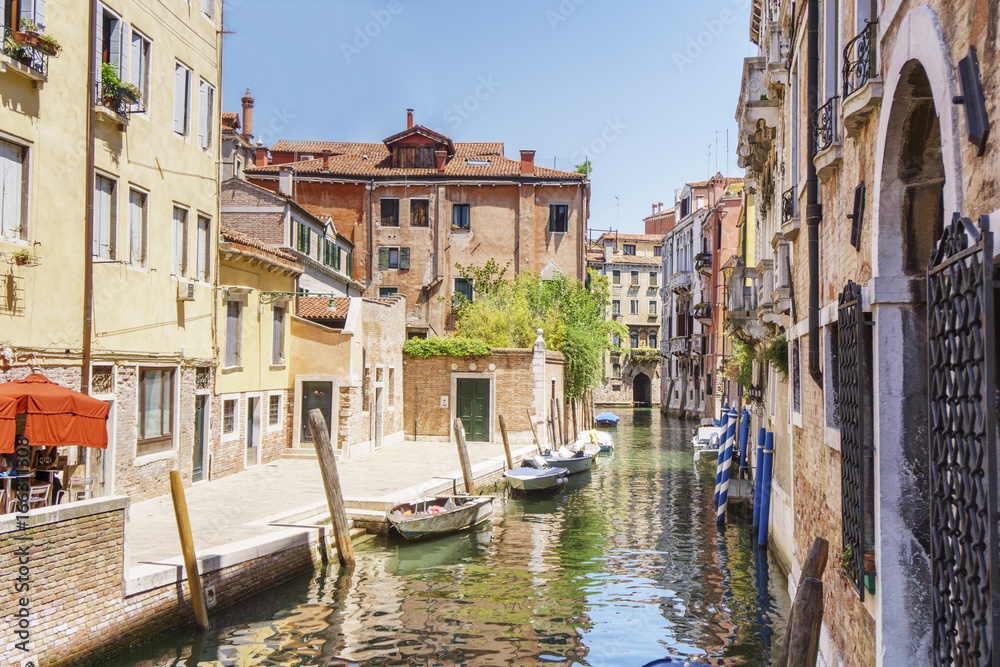 Venice as it is: canal, gondolas, bright buildings and tourists at noon time.