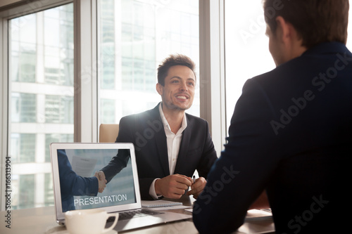 Smiling investment broker showing digital presentation on laptop screen to client, friendly salesman giving sales pitch, making business proposal to partner, selling product, offering cooperation